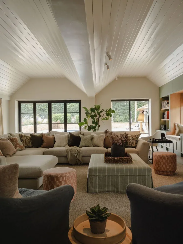 A photograph of a comfortable living room with a white vaulted ceiling and large windows. It features an L-shaped sofa, a striped ottoman, and a fiddle leaf fig plant. Two armchairs are in the foreground.