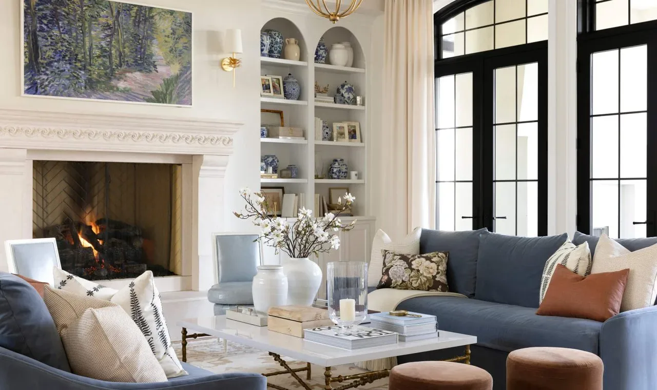 Elegant Blue and Cream Living Room with Classic Fireplace Designed by Decozie Designer Kate C. Refined living room featuring blue sofas, cream accents, a classic white fireplace with a burning fire, built-in shelving with decorative ceramics, and black-framed French doors, designed by Decozie designer Kate C.