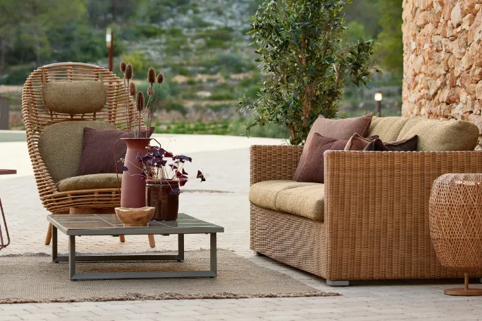 Curated outdoor living space on a patio, featuring a high-backed natural rattan cocoon chair and matching two-seater wicker sofa with olive green and brown cushions, on a natural jute rug. Background of a textured stone wall and Mediterranean hillside.
