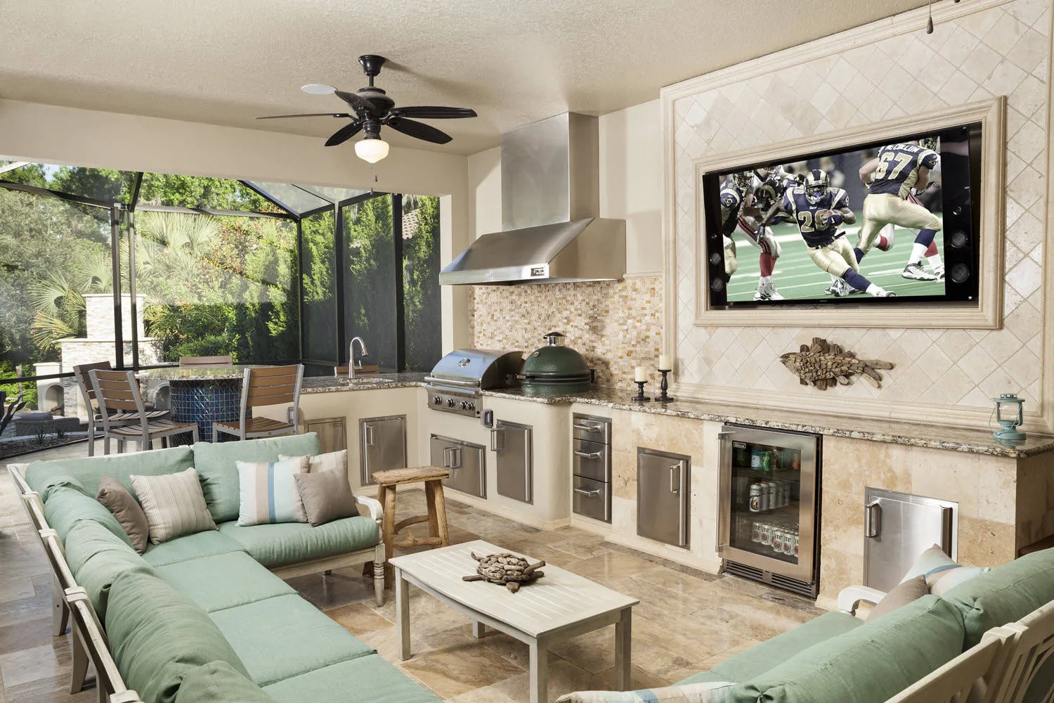 A professional-grade outdoor living and entertainment suite featuring a complete outdoor kitchen with stainless steel appliances and a built-in smoker. The space is expertly zoned for relaxation with a spacious sage green sectional sofa lounge and a wall-mounted outdoor television. High-quality finishes, including granite countertops and tiled walls, create a designer-level transformation. In the background, a screened-in dining area illustrates efficient space optimization for year-round use.