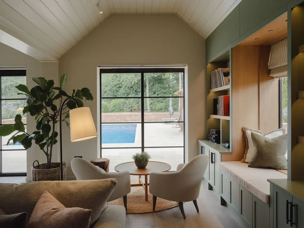 Cozy modern living room with beige sofas, armchairs, floor lamp, built-in window seating, and indoor-outdoor pool view