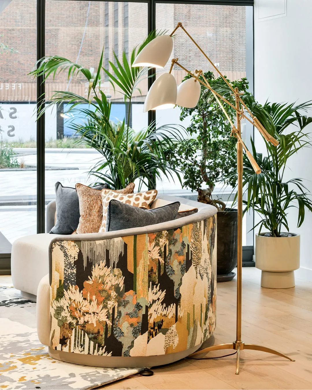 Curved Patterned Sofa and Modern Interior Decor A bright interior corner featuring a curved modern sofa with an abstract nature-patterned back, a brass three-armed floor lamp, and large potted plants.
